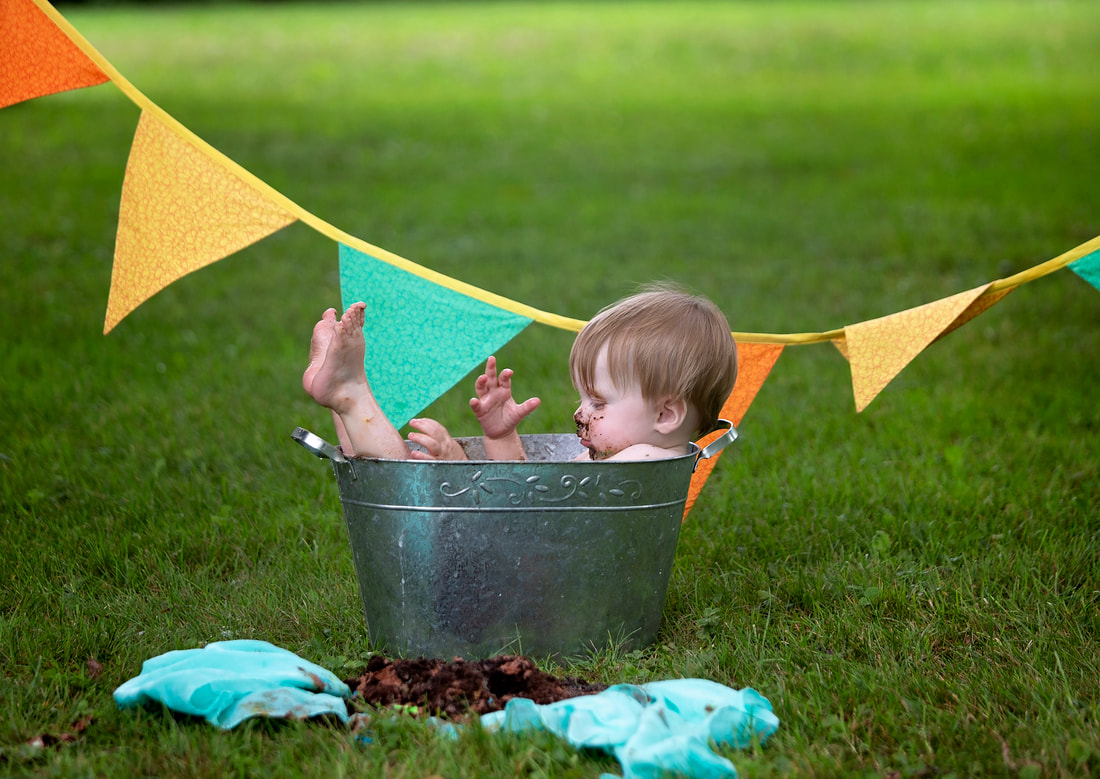 Photo of a baby in a small tin bathtub on the grass after smashing his cake with flag banner and cake all around him. Cake smash photo session