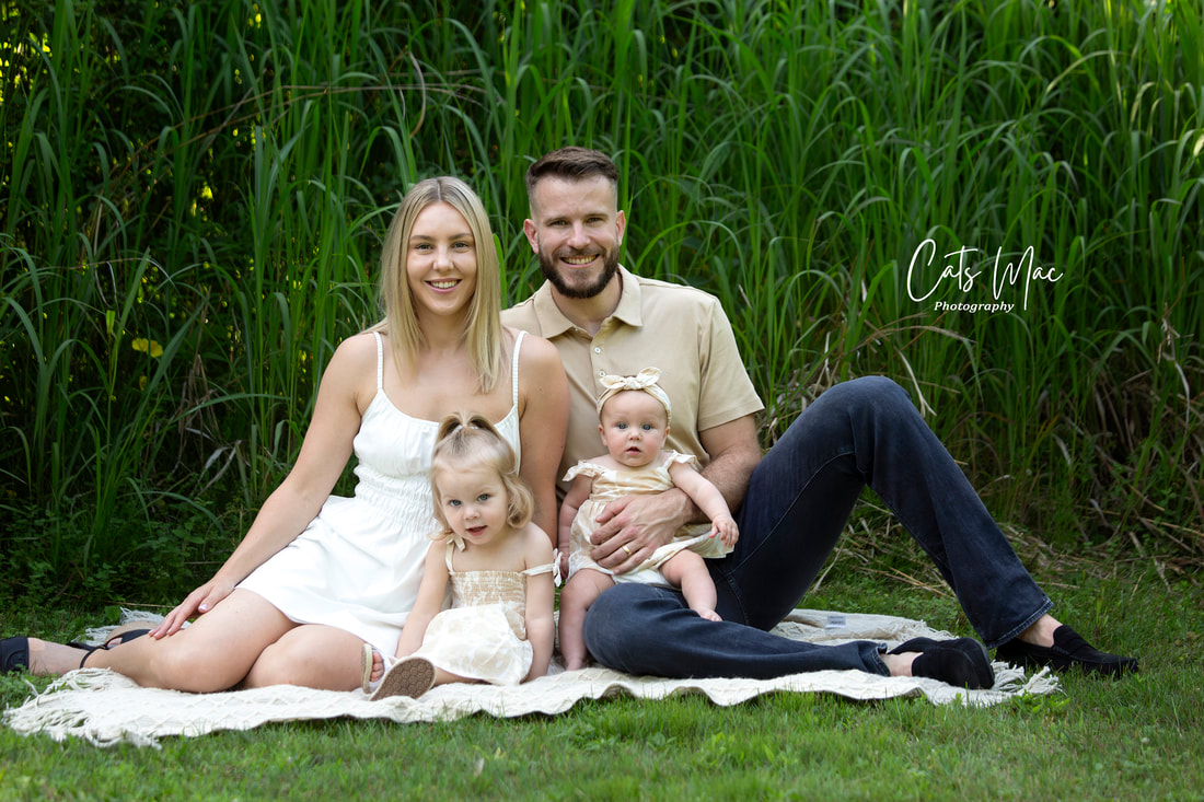 Mom dad girl and baby sitting on grass against tall bush outside family photo session 