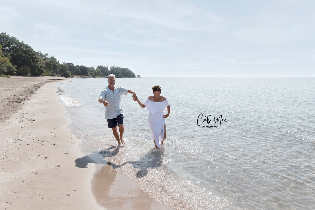 Grandparents dressed in white holding hands and running along the shoreline of Cobourg Beach 