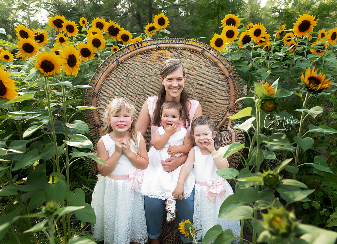 Mom and three little girls sitting in a peacock chair among a large sunflower patch