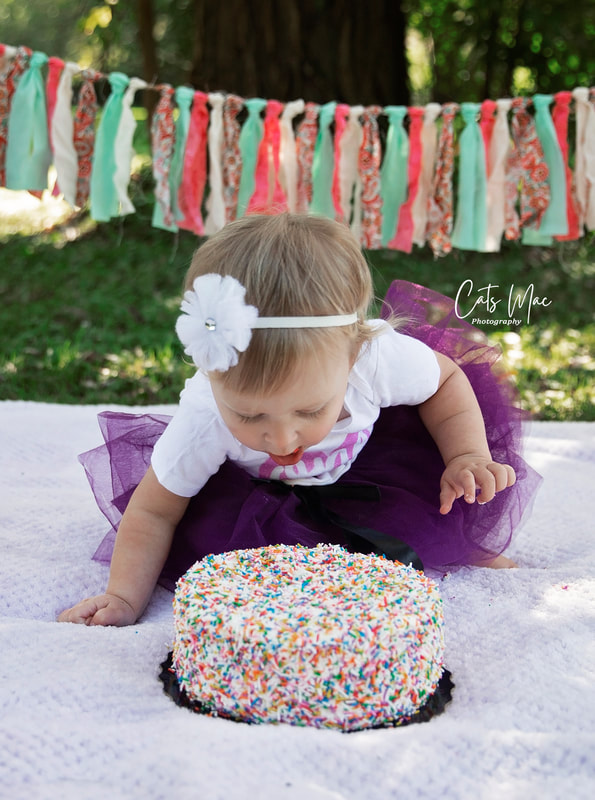 Baby girl dressing in purple tutu bending down towards her birthday cake with her mouth open