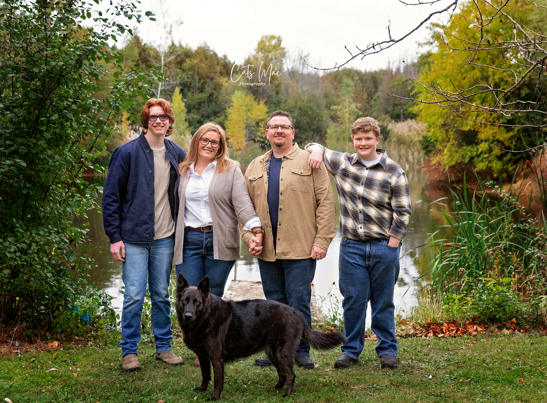 Mom, Dad and two brothers posing with their black dog in front of pond fall colours photo shoot