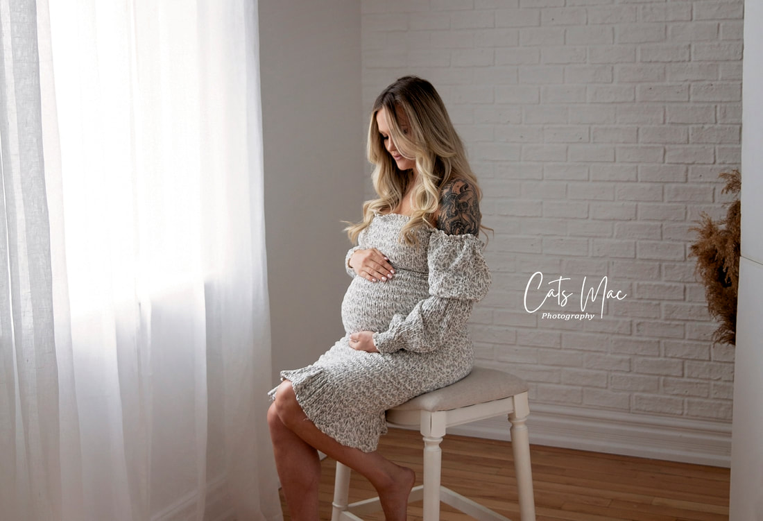 Maternity Photo Session pregnancy woman sitting on stool by natural light from a curtained window looking down at her belly